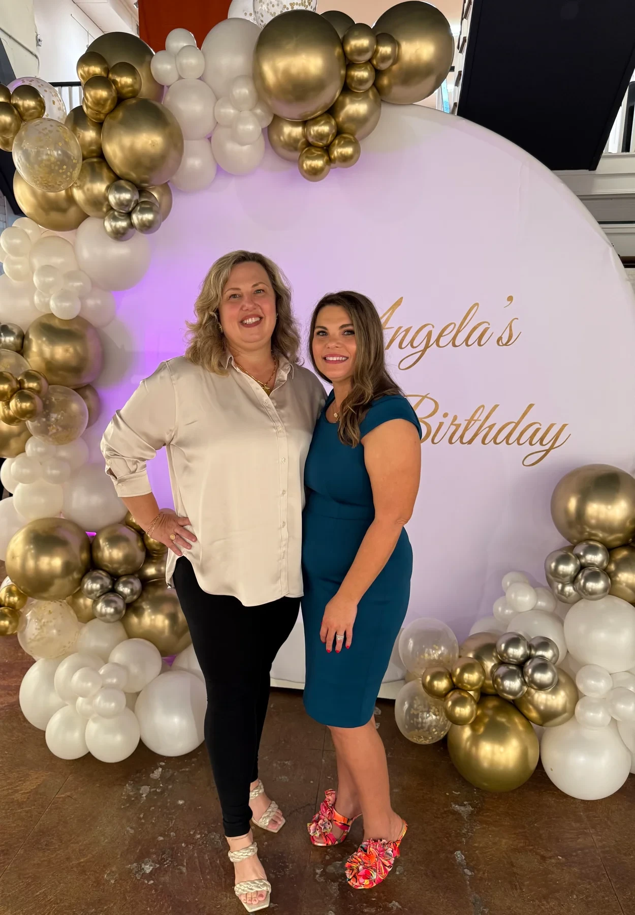 Two women posing together beneath the gold and white balloons.