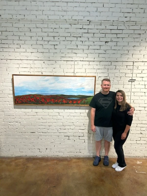 Two people posing beside a long landscape painting of red flowers.